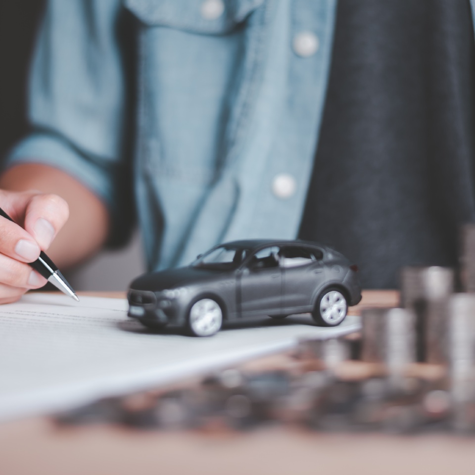 A person writing on paper next to a small black toy car and stacked coins, suggesting financial planning or car insurance.