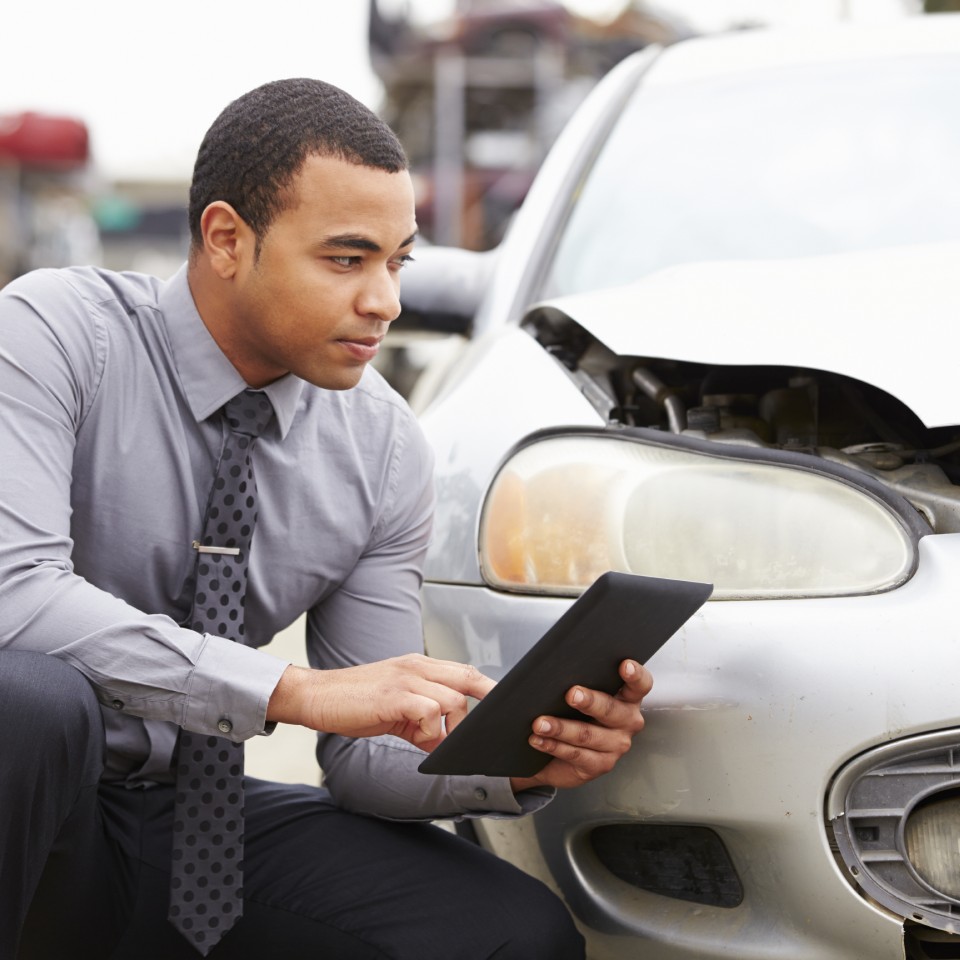 Man in business attire inspects a damaged car and takes notes on a tablet, focusing on the crumpled front part of the vehicle.