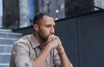 A man in a beige shirt sits with his hands clasped in front of his face, appearing deep in thought or concerned, with steps and a gray wall in the background.