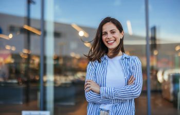 A woman stands outdoors in front of a glass building, smiling with her arms crossed, wearing a white shirt and a blue-and-white striped overshirt.
