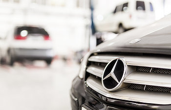 Close-up of a Mercedes-Benz car grille with the logo visible; blurred vehicles and a garage setting in the background.