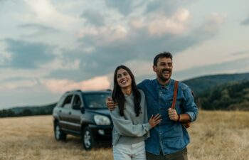 Loving couple embracing and following the trail along a grassy mountain ridge and having fun together. Car in the background